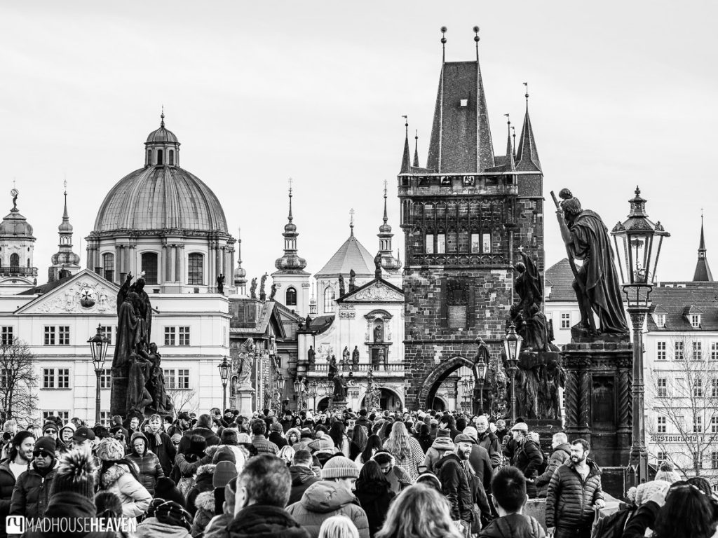 Black and white image of the Charles Bridge, with many people, statues, domes and towers of Prague in it